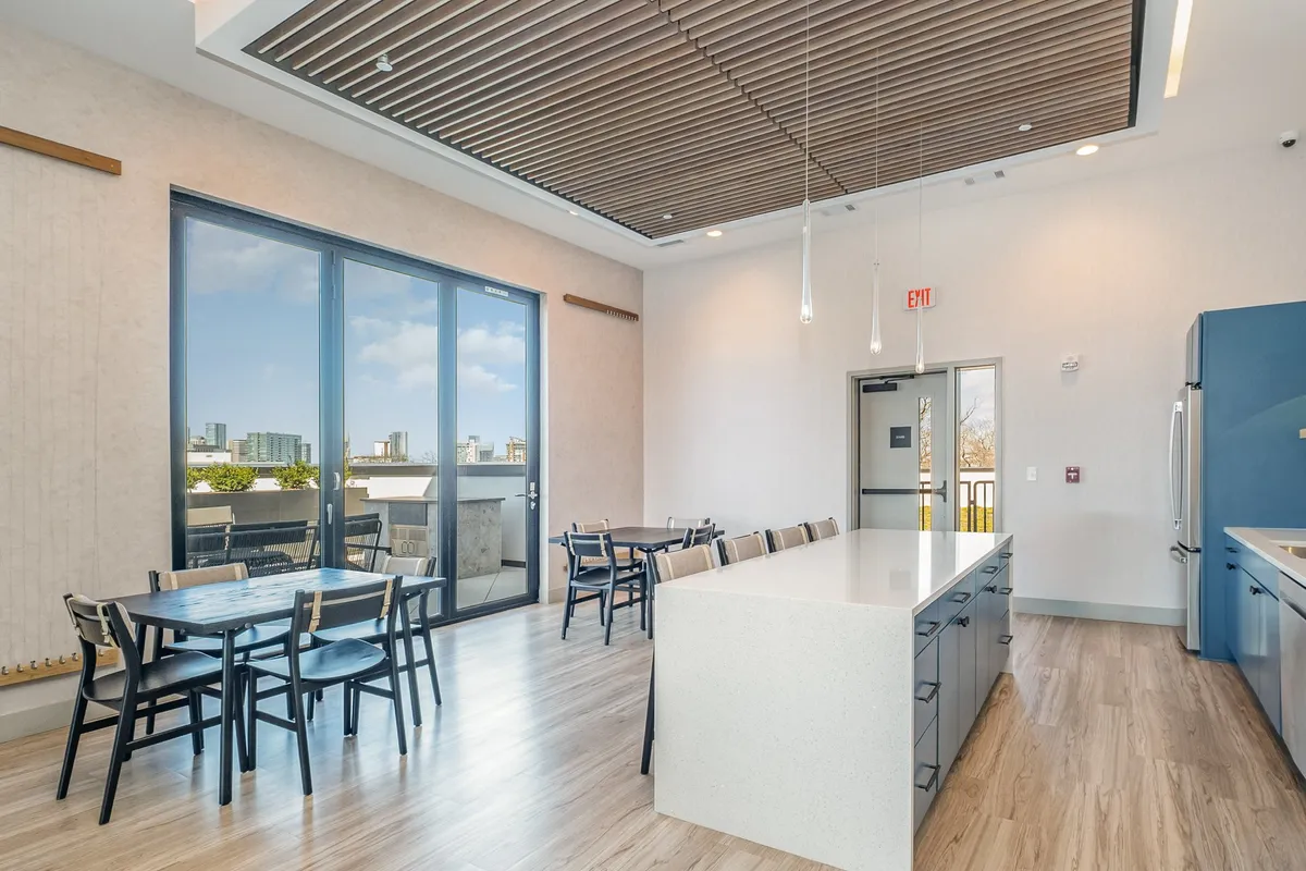 An open-plan interior featuring large windows, a spacious kitchen island, and dining tables arranged for group meals. The modern design and natural light suggest a seamless connection between kitchen and dining areas—perfect for gathering and sharing meals.