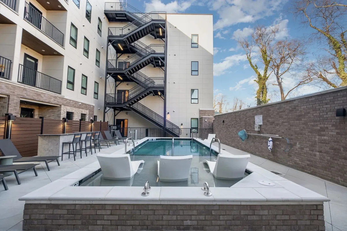 This image depicts a sparkling swimming pool surrounded by lounge chairs and an outdoor seating area, set against a backdrop of modern residential buildings. The pool area feels inviting, perfect for both swimming and sunbathing, with space for socializing and relaxing outdoors. The overall atmosphere is leisurely and relaxed, capturing the essence of resort-style amenities within an urban environment.