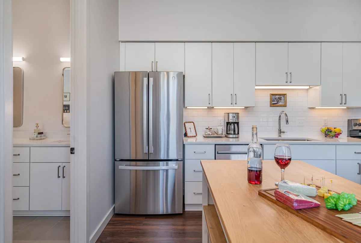 This image showcases a bright, modern kitchen featuring a stainless steel refrigerator, a spacious wooden kitchen island, wine glasses, and a colorful cheese platter. The space is clearly set up for both cooking and entertaining, with inviting finishes and social touches.