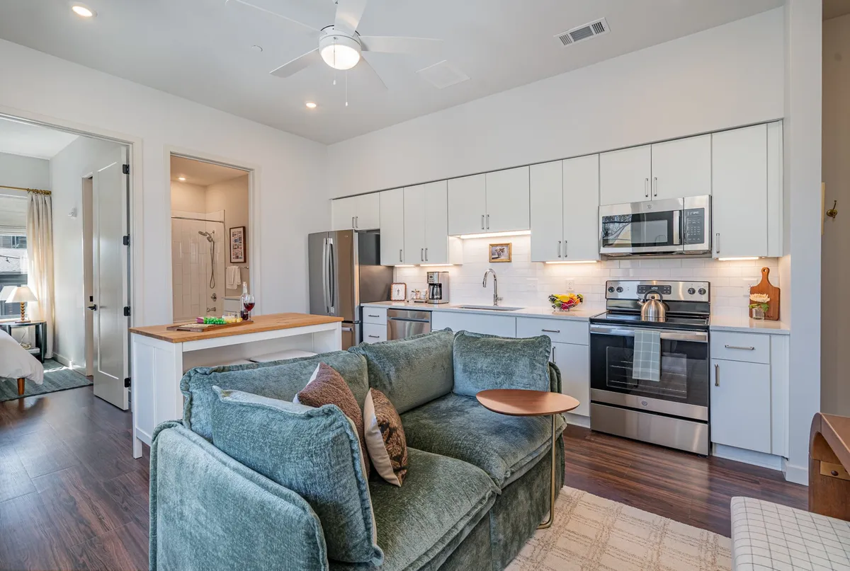 A bright, open-concept living area featuring a plush sectional sofa, a spacious island counter, and gleaming stainless steel kitchen appliances. The space is styled with modern touches and natural light, ideal for gathering, cooking, and relaxing.