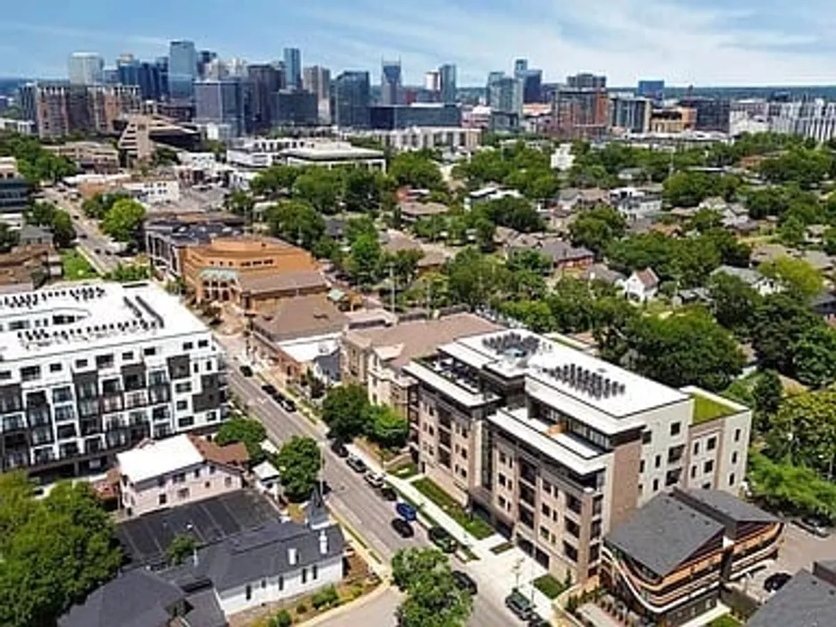 A vibrant urban exterior showing modern apartment buildings, Nashville’s downtown skyline, green spaces, and tree-lined streets. The scene is lively, with people strolling and gathering, suggesting the area’s energy and accessibility to parks and local events.