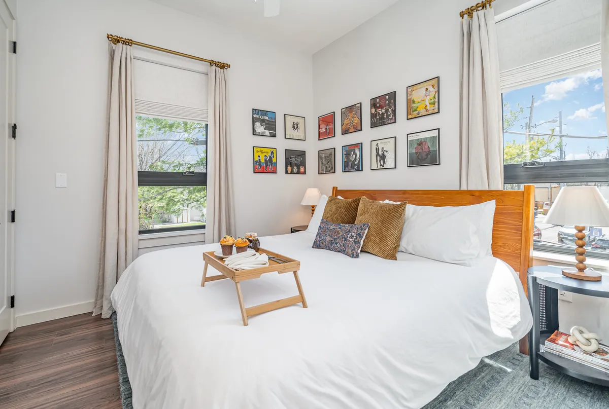 A tranquil bedroom with a plush bed adorned with decorative pillows, a breakfast tray with coffee and pastries, sunlight streaming through the window, and framed artwork providing a serene, cozy atmosphere.