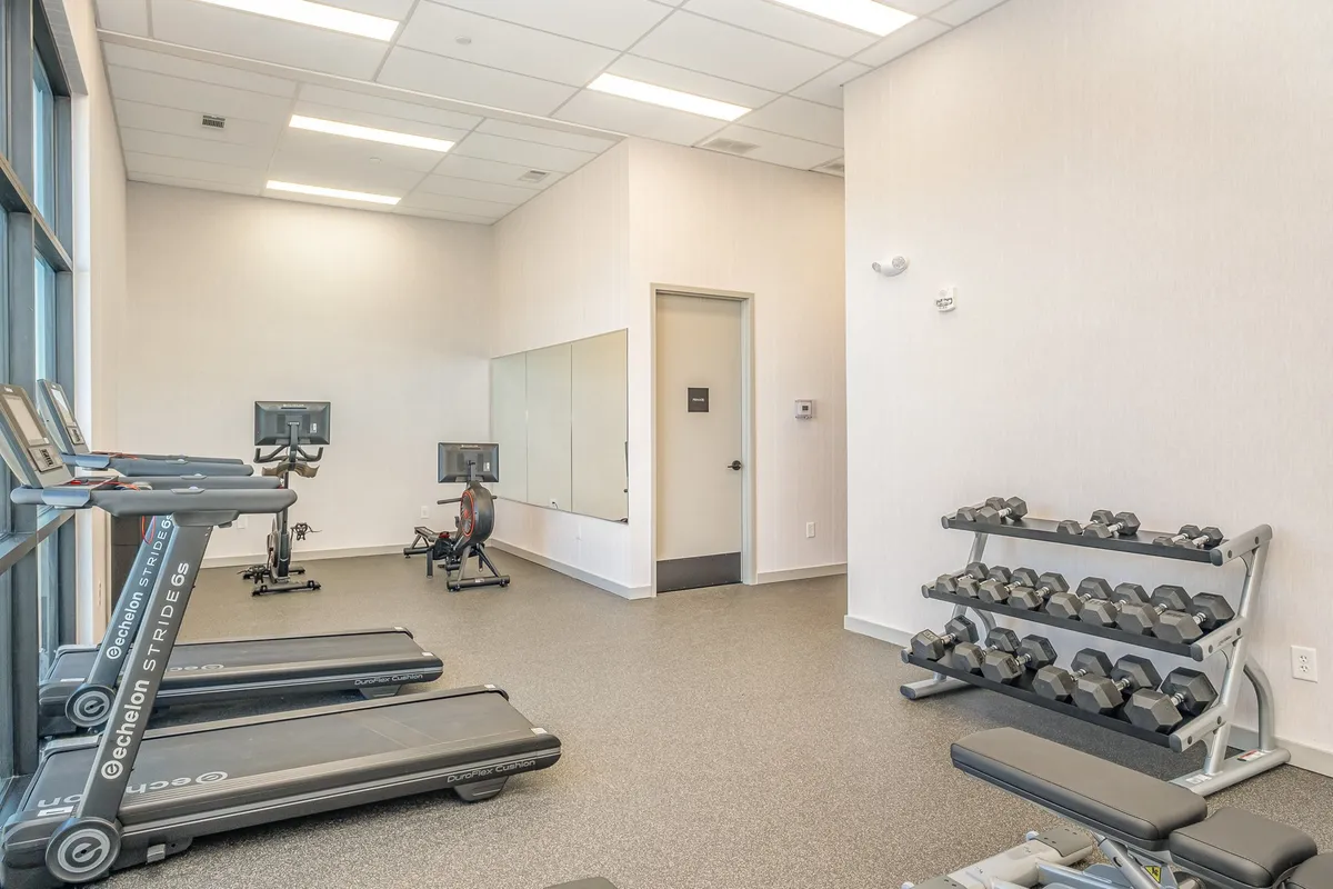 This interior image showcases a well-equipped gym featuring treadmills, a dumbbell rack, an exercise bike, and large mirrors. The space is bright and invigorating, ideal for maintaining fitness routines and participating in group workouts. The gym setting feels modern and motivating, appealing to wellness-focused residents.